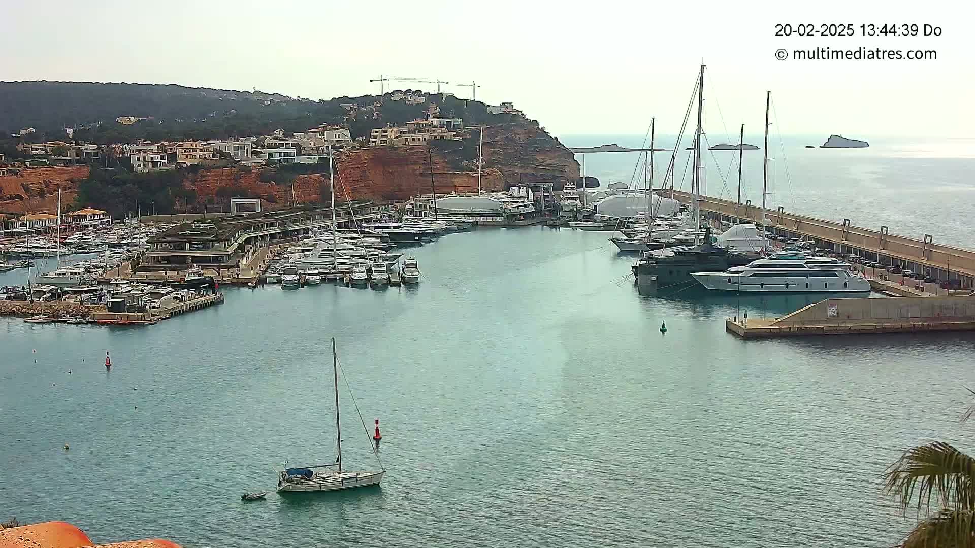 A calm, overcast day reveals a marina filled with numerous yachts and sailboats, nestled against a rocky coastline dotted with buildings.