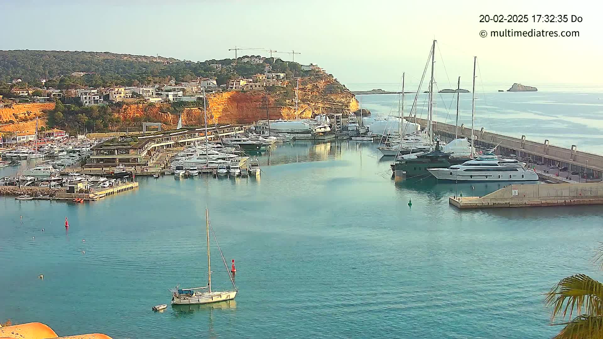 A marina filled with yachts and sailboats is situated near a hillside town under a clear sky.