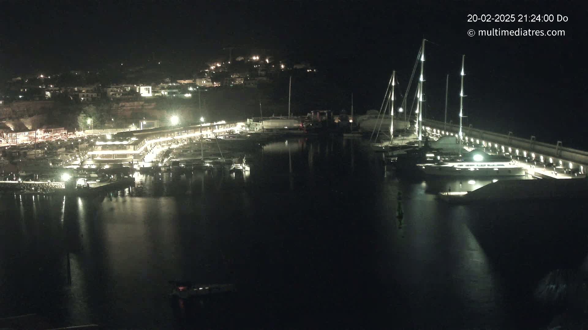 A nighttime view of a harbor filled with boats and yachts, illuminated by artificial lights, with a town visible on a hill in the background.
