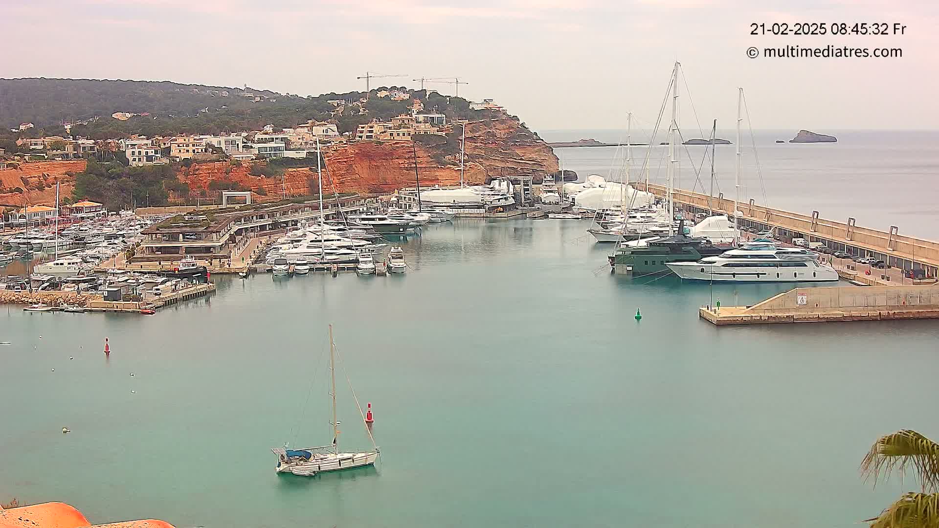 A marina filled with numerous yachts and sailboats is nestled against a red-cliff hillside under an overcast sky.