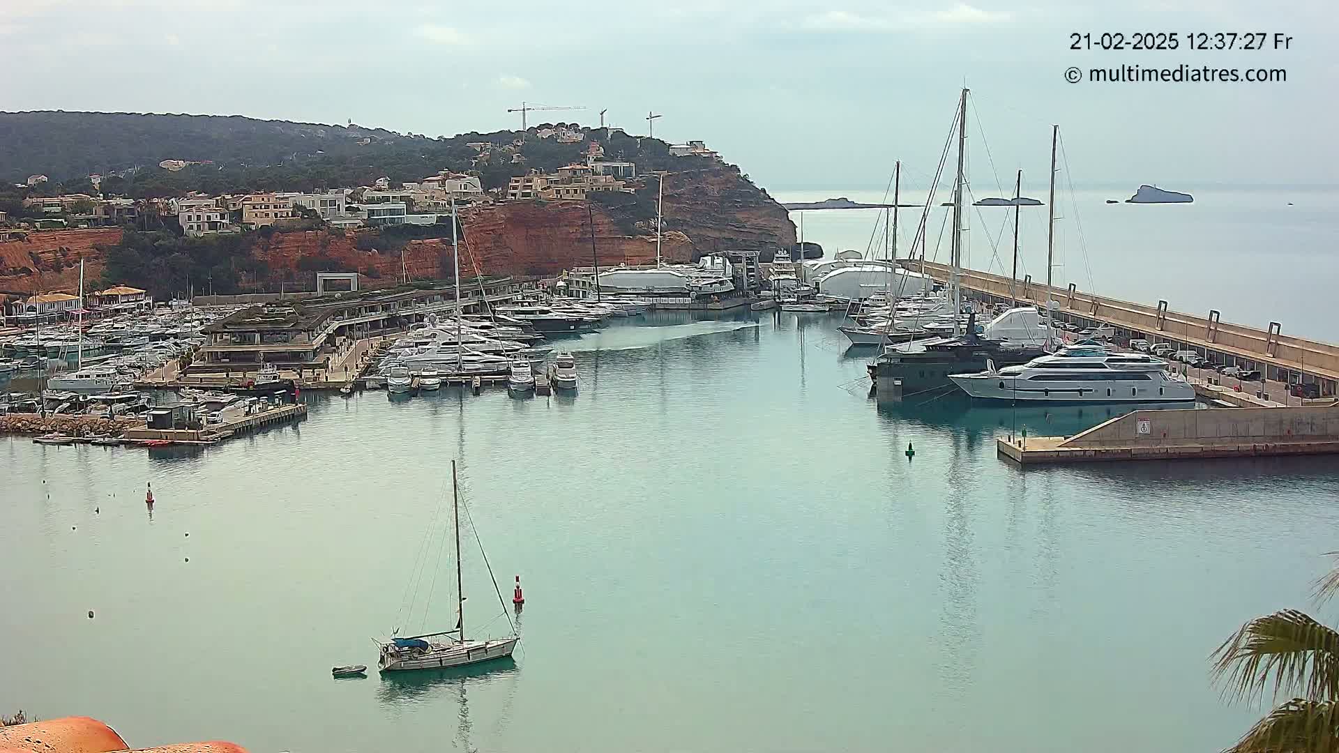 A marina filled with numerous yachts and sailboats is situated in calm, clear water next to a hillside town under an overcast sky.