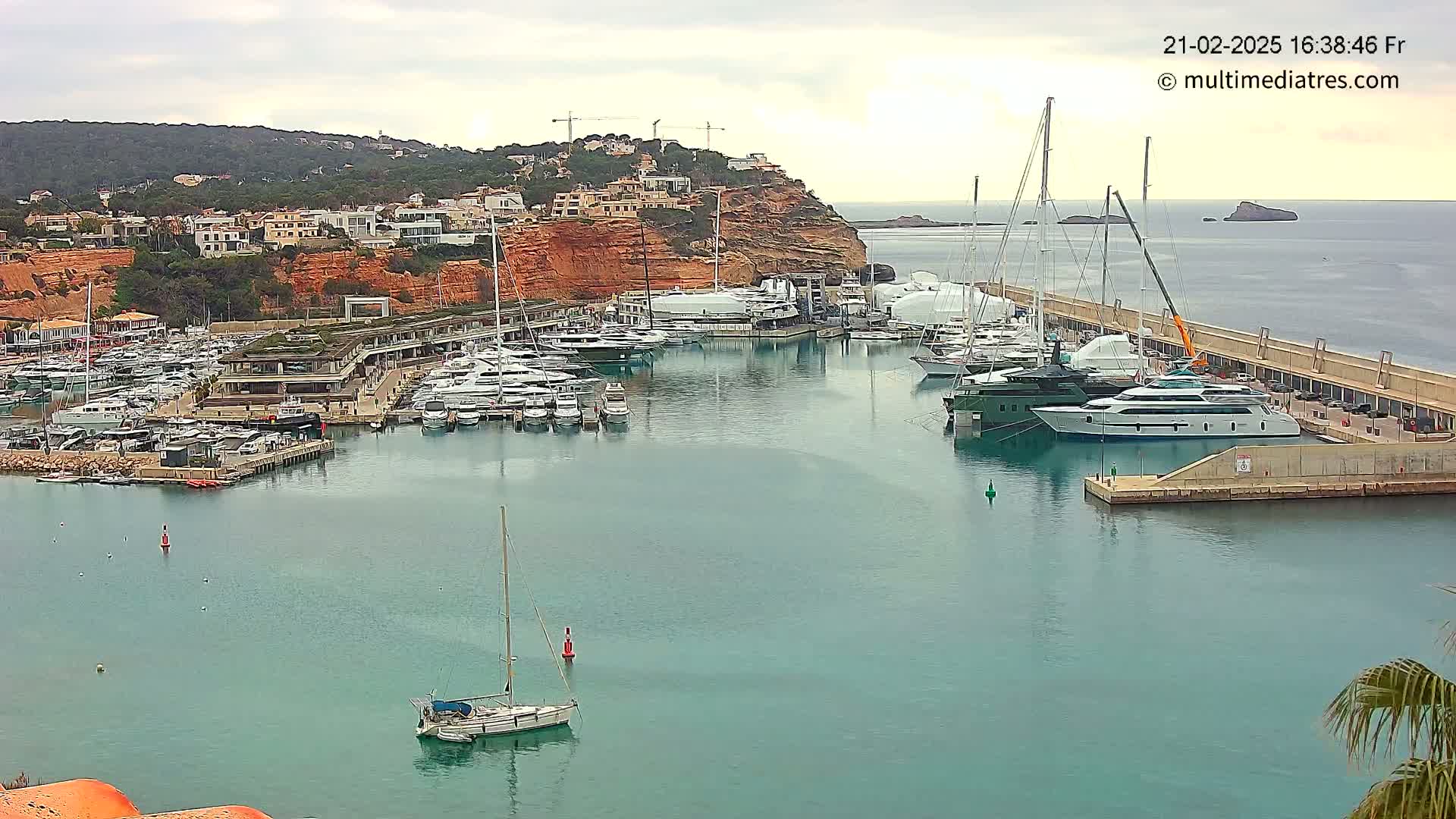 A harbor filled with numerous yachts and sailboats is nestled against a hillside of reddish-brown cliffs under an overcast sky.