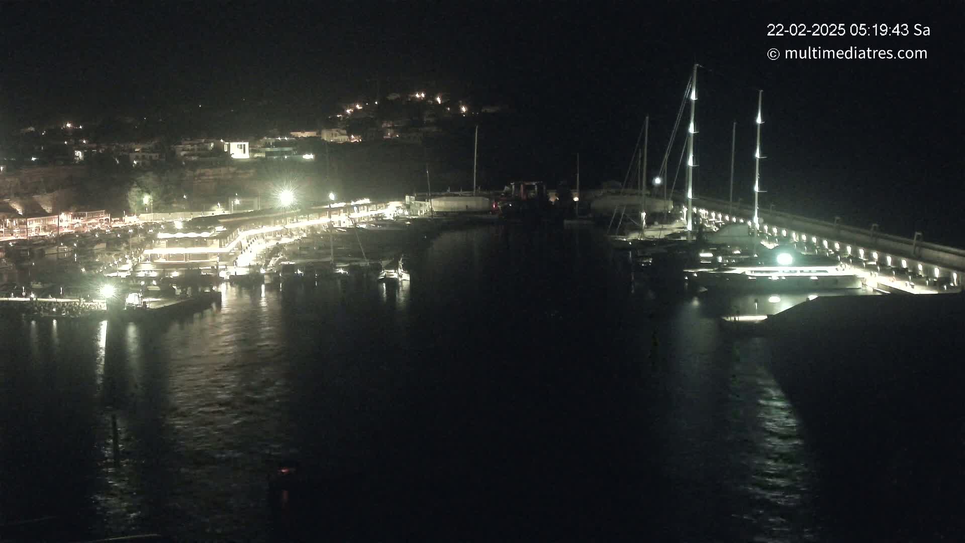 A nighttime view of a marina filled with boats and yachts, illuminated by lights along a dock and buildings on the shore.