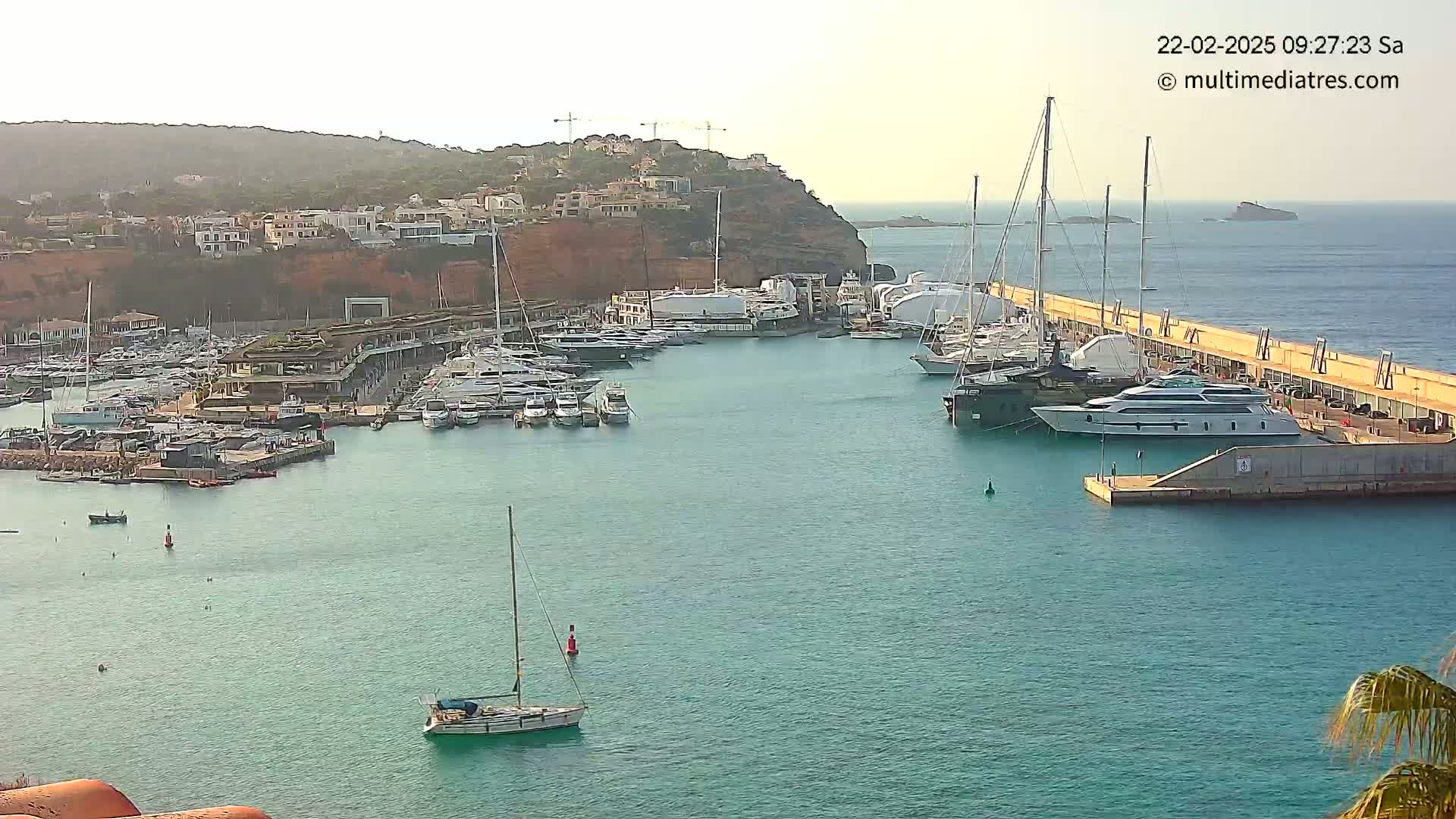 A marina filled with yachts and sailboats is situated in a calm, turquoise body of water near a reddish-brown cliffside with buildings on top.