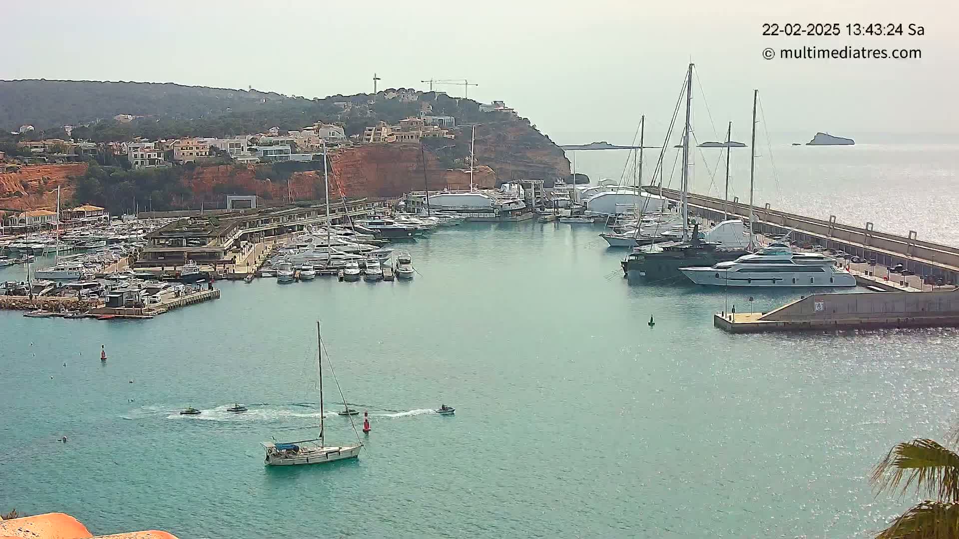 A marina filled with numerous yachts and sailboats is situated on calm, turquoise water next to a red-cliffed hillside under an overcast sky.