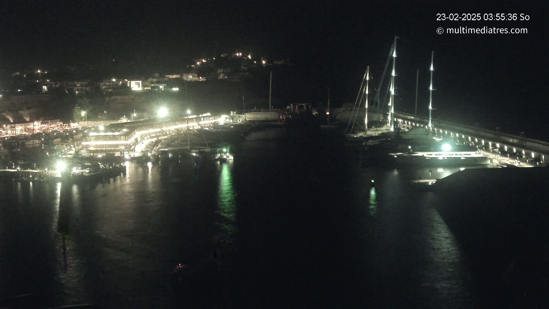 A nighttime view of a harbor with boats docked, buildings lit up along the shoreline, and several tall masts of sailboats visible against a dark sky.