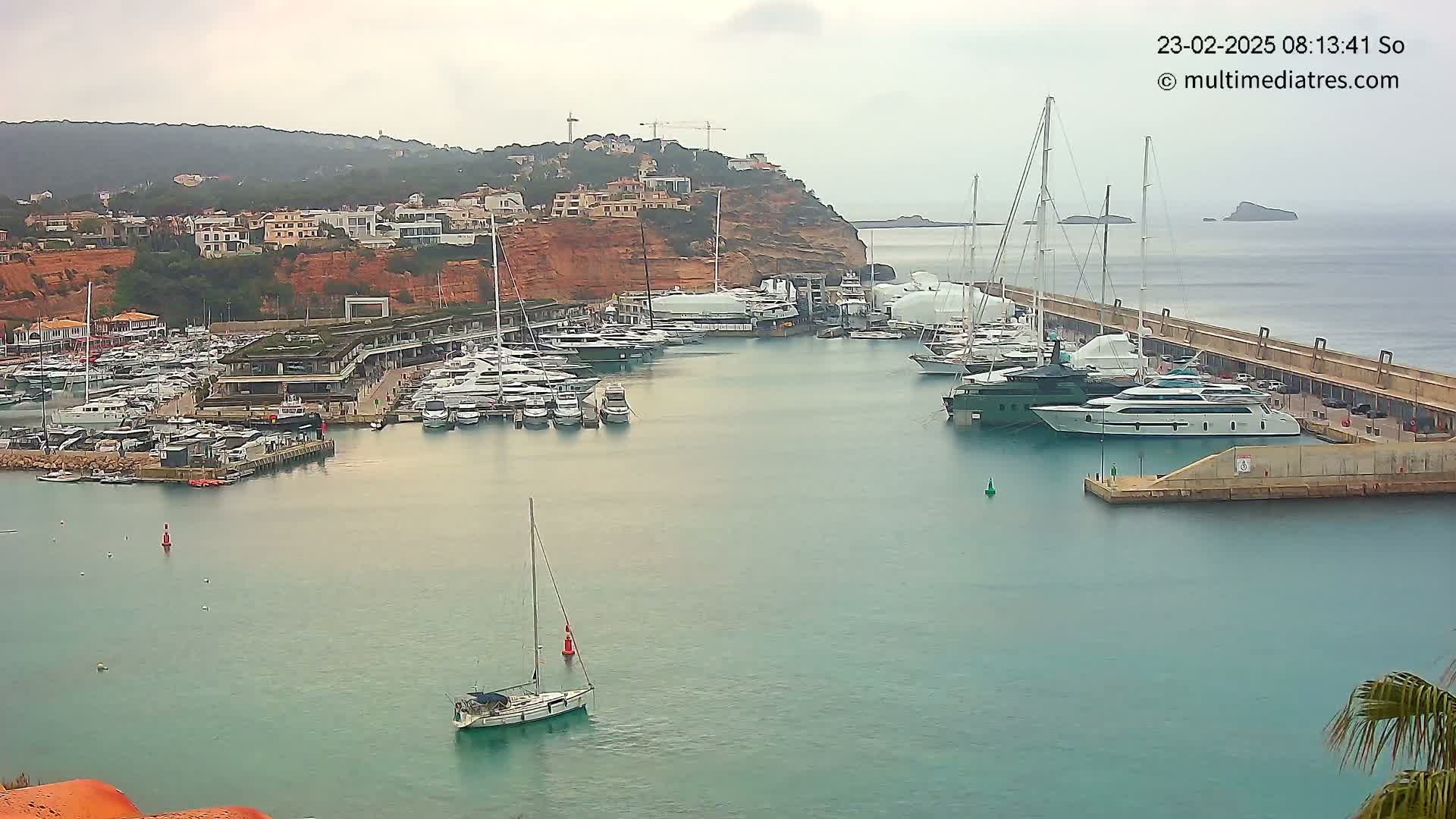A marina filled with numerous yachts and sailboats is situated on calm, teal water next to a hillside town under an overcast sky.