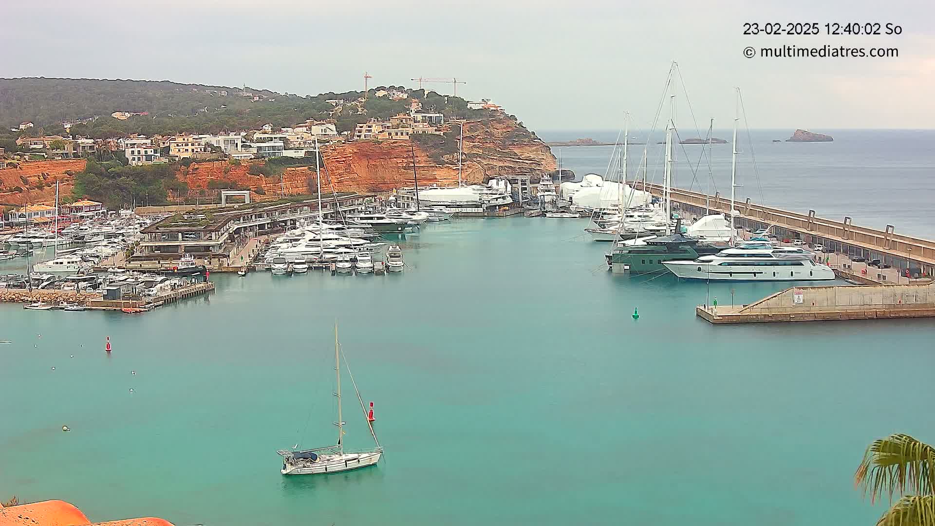 A marina filled with numerous yachts and sailboats is nestled against a red-clay hillside under an overcast sky.