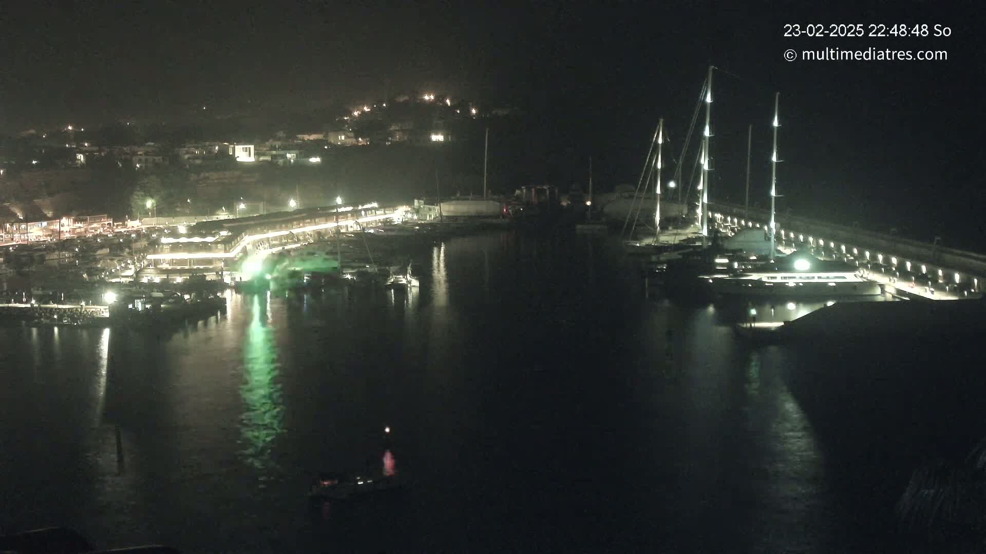 A nighttime view of a marina filled with boats, illuminated by lights reflecting on the calm water, with a town visible in the background on a hill.