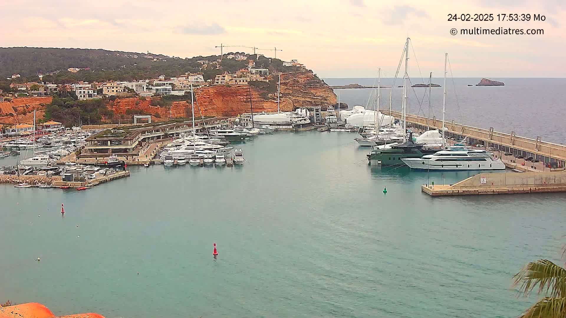 A calm harbor filled with numerous yachts and boats is nestled against a red-cliffed coast under an overcast sky.