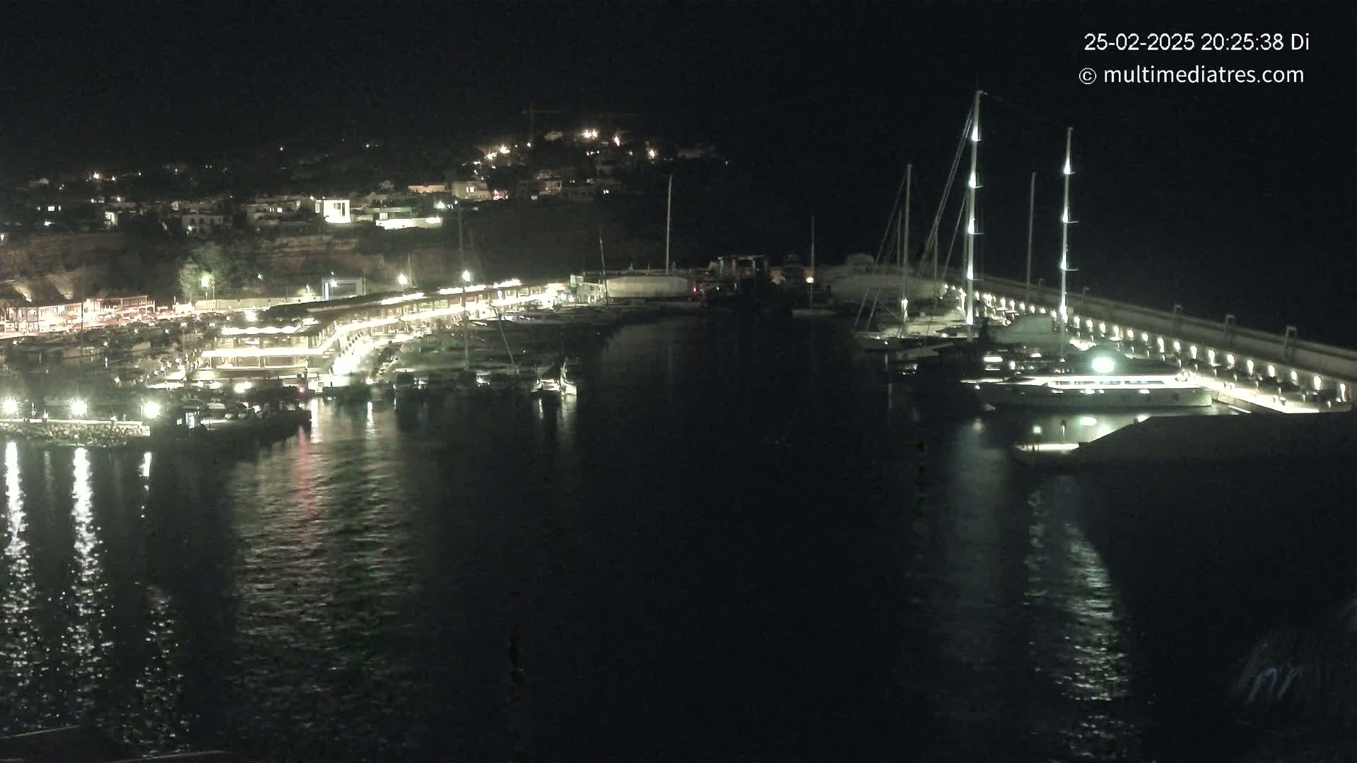 A nighttime view of a marina filled with boats, illuminated by lights along a waterfront with buildings on a hillside in the background.