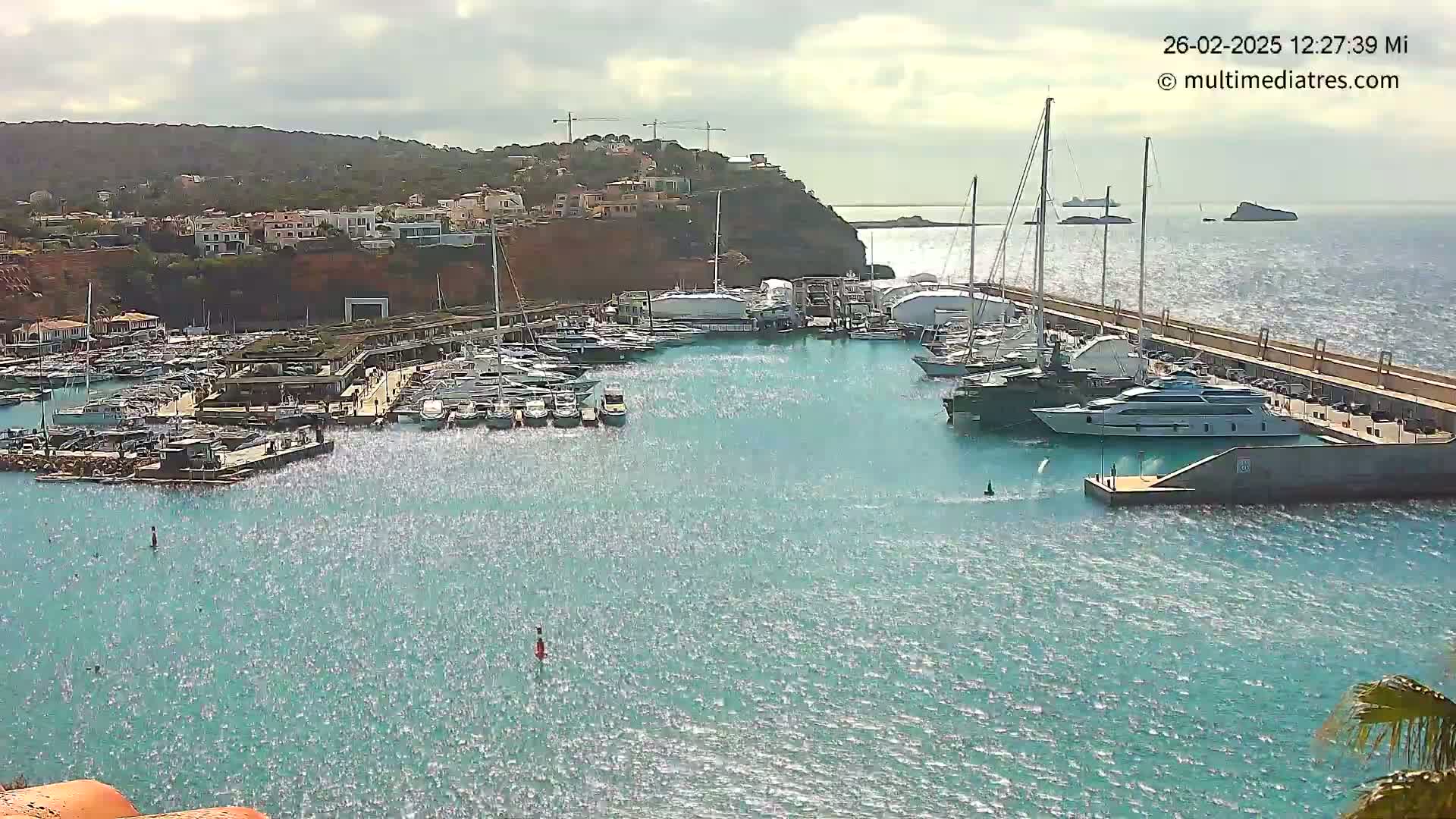 A marina filled with numerous yachts and boats is situated on a calm, sparkling body of water next to a hillside town under a partly cloudy sky.