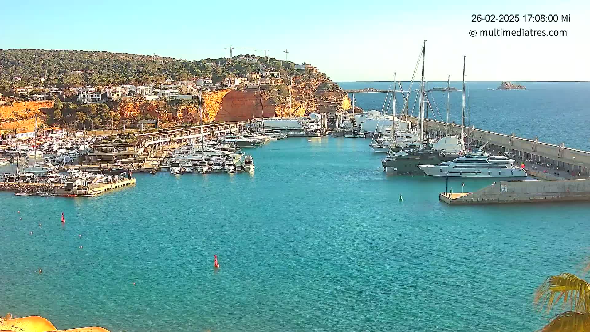 A marina filled with numerous yachts and boats is situated against a backdrop of a sunny, hilly coastline.