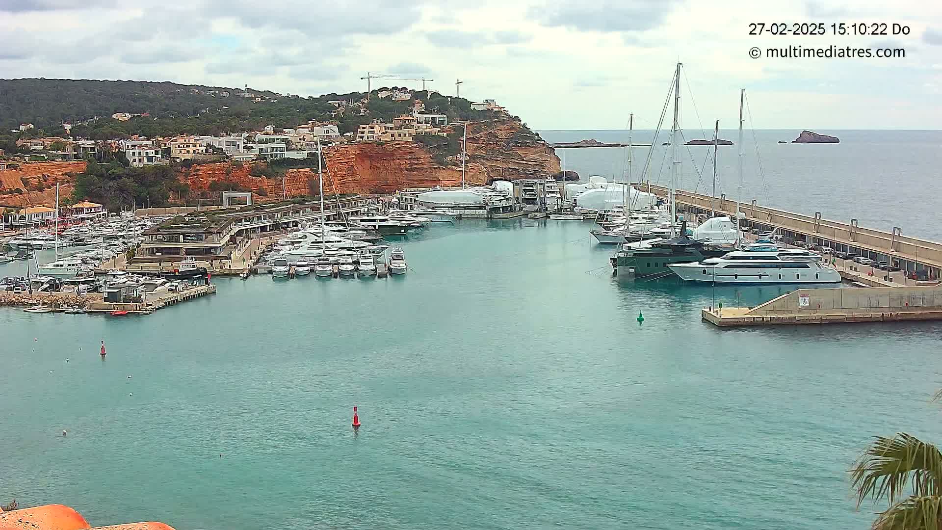 A marina filled with yachts and boats is nestled against a hillside of red-toned cliffs under an overcast sky.