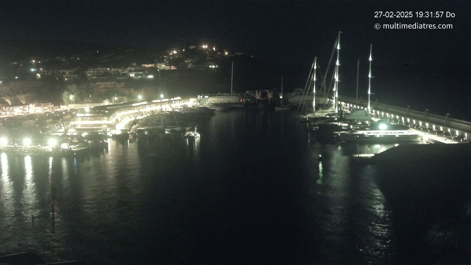 A nighttime view of a marina filled with boats and yachts, illuminated by lights, with calm water reflecting the lights.