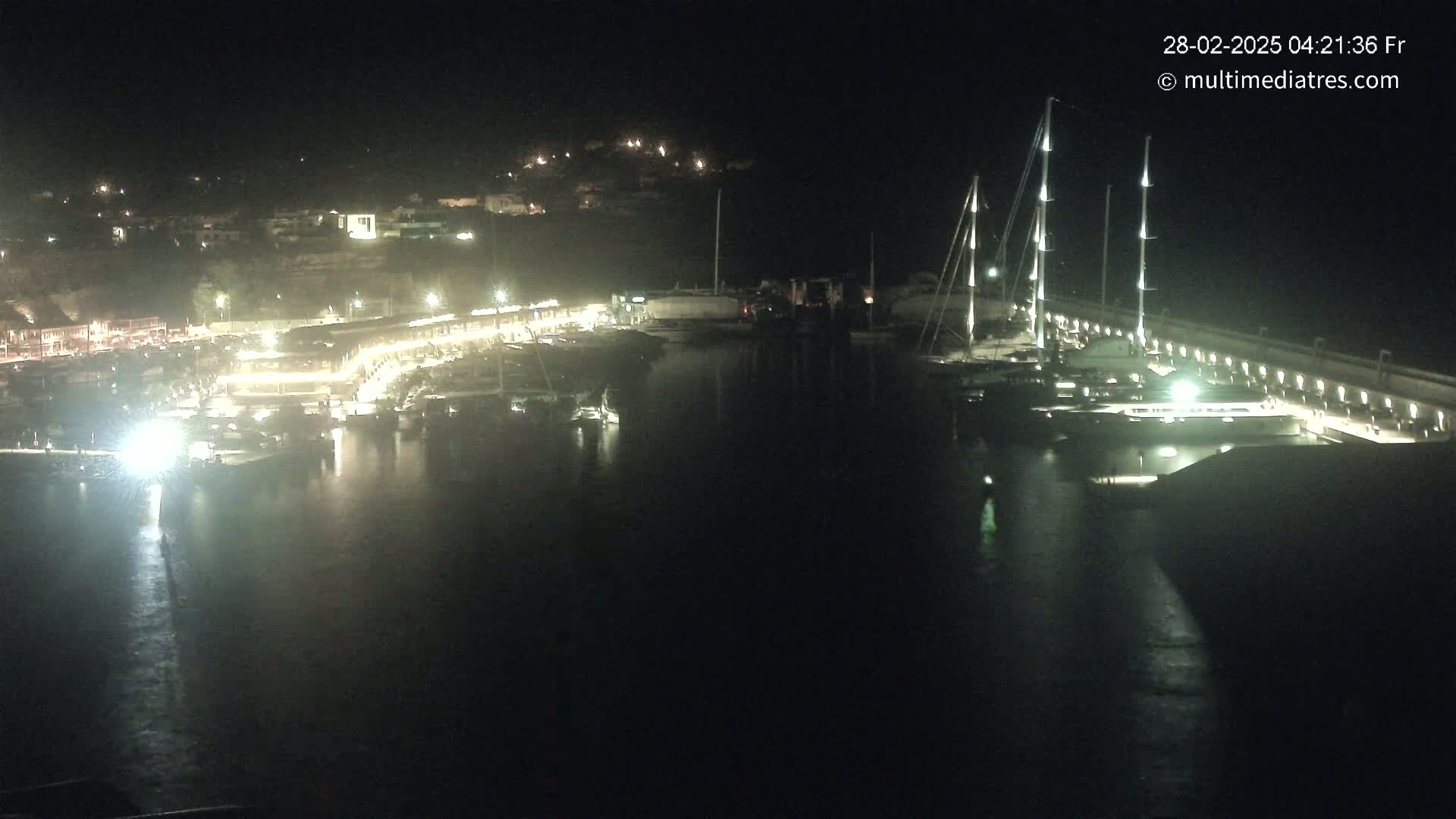 A nighttime view of a marina, illuminated by lights, with several boats docked and a town visible in the background.
