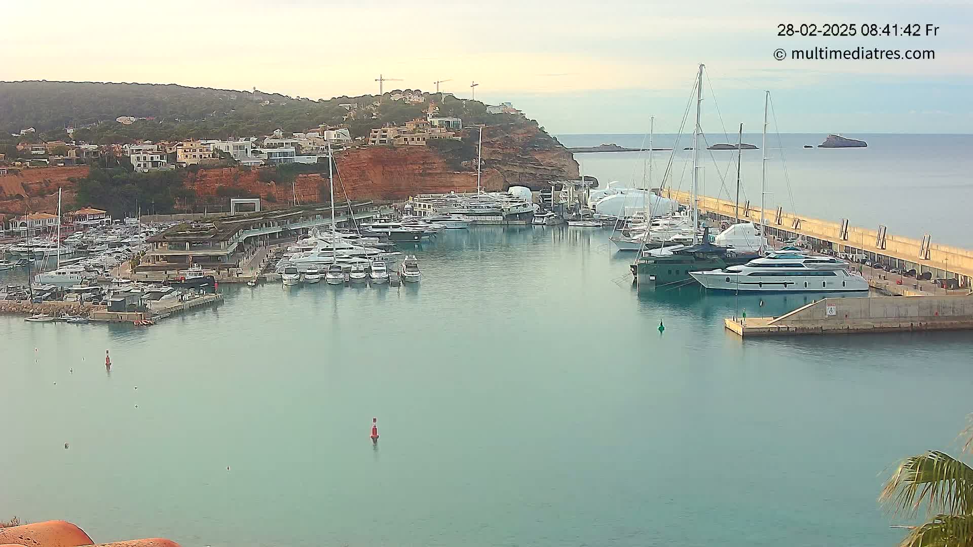 A marina filled with many yachts and sailboats is nestled in a calm bay next to a red cliffside with buildings on top under a mostly clear sky.