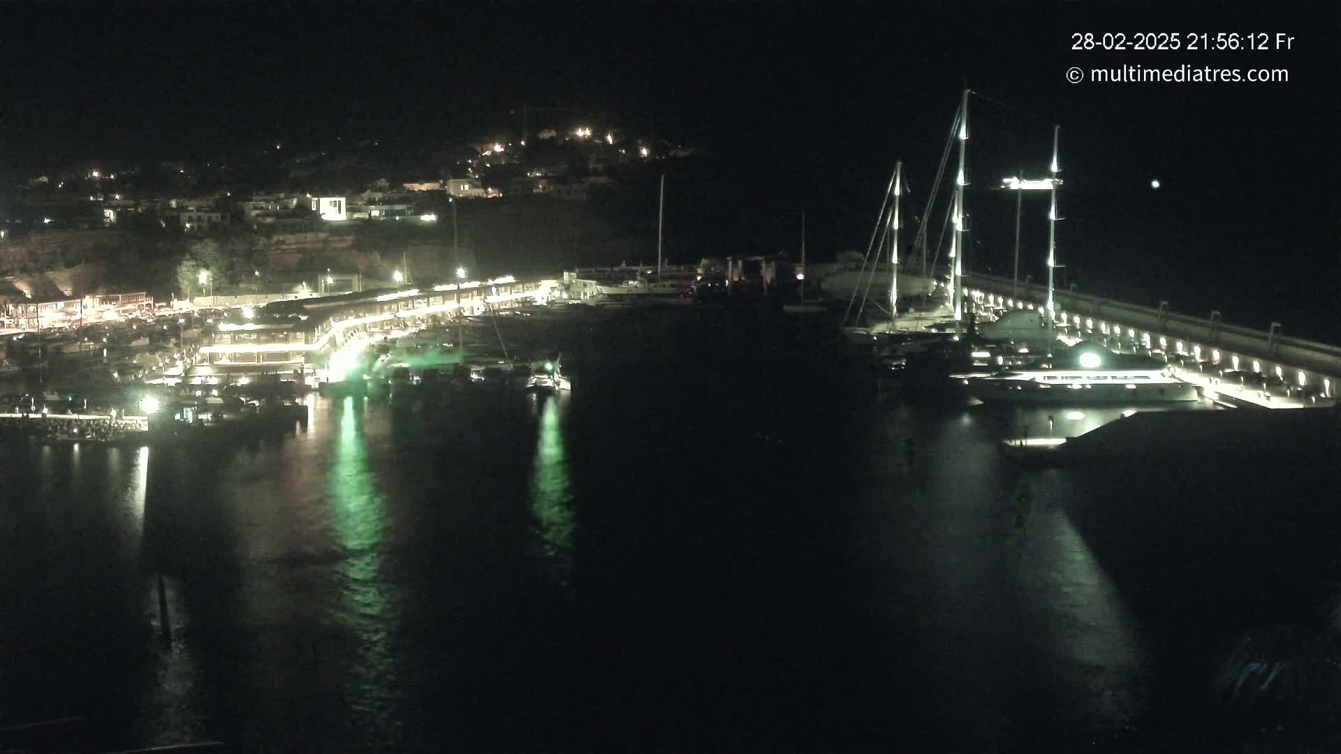 A nighttime view of a marina filled with boats, illuminated buildings along the shoreline, and calm water reflecting the lights.