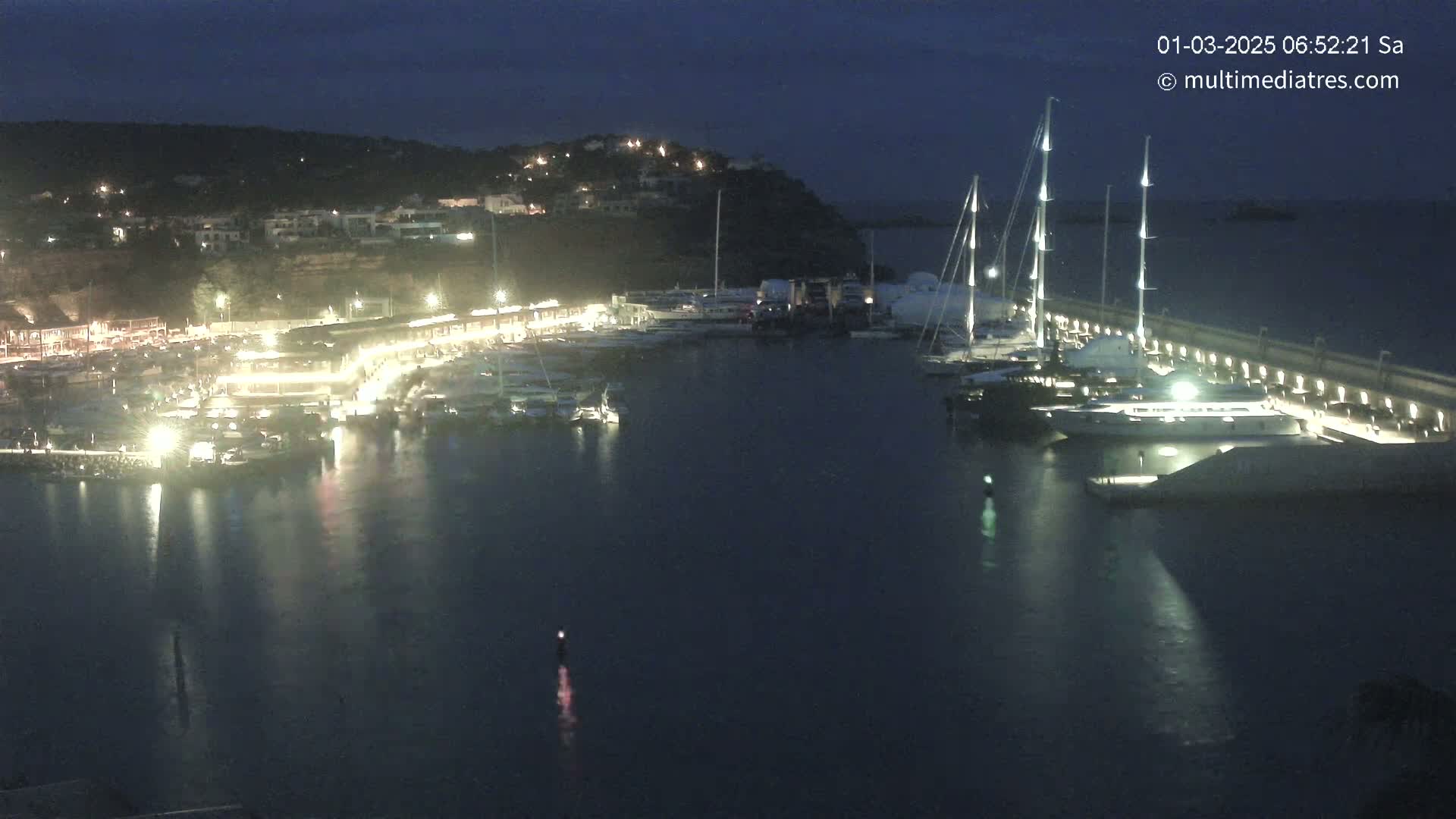 A nighttime view of a harbor filled with boats docked along a lit waterfront, under a dark sky.