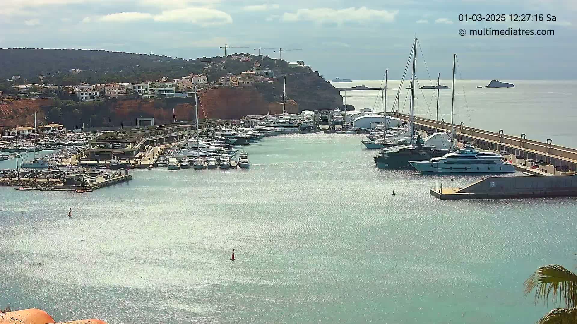 A marina filled with numerous yachts and sailboats is situated next to a red cliff housing multiple buildings, under a partly cloudy sky.