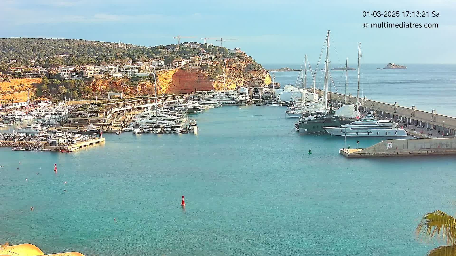 A marina filled with yachts and boats is situated in a calm, clear body of water next to a hillside of buildings under a sunny sky.