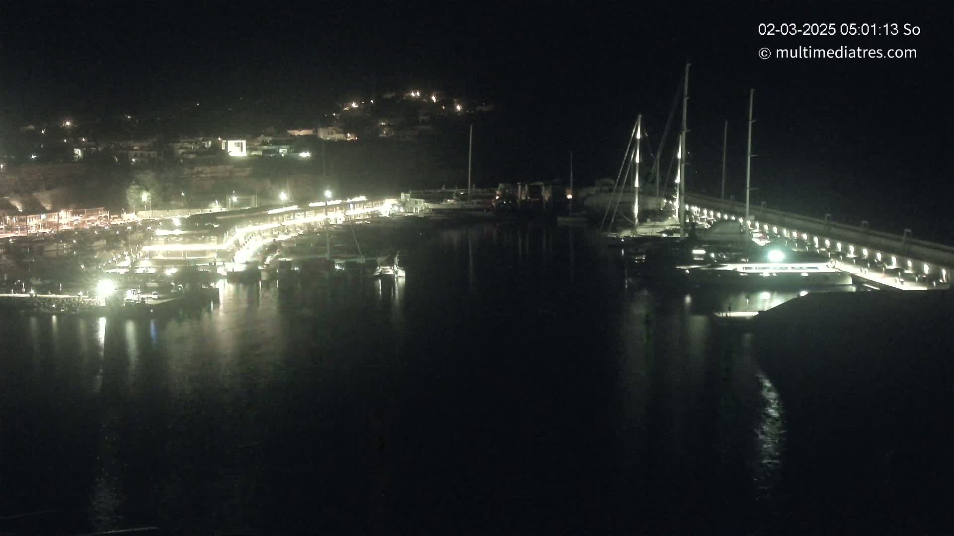 A nighttime view of a harbor filled with boats, illuminated buildings lining the shore, and calm water reflecting the lights.