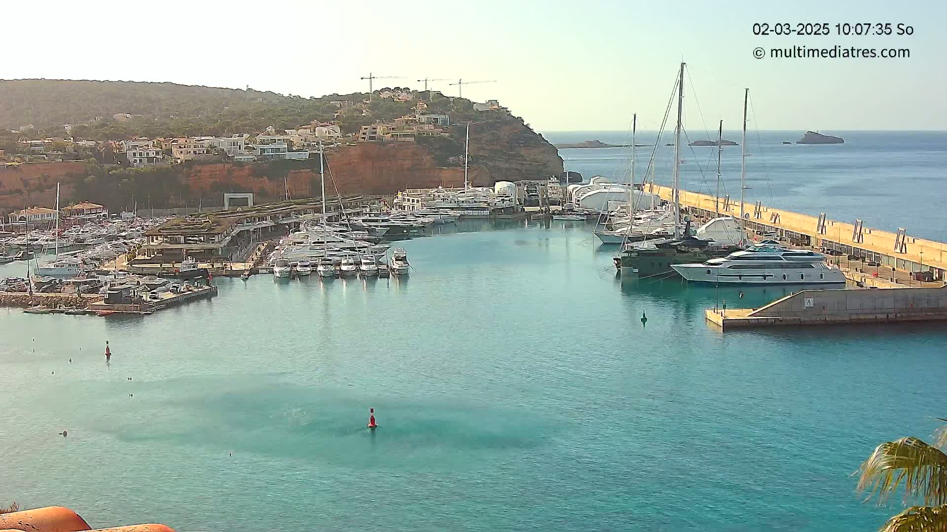 A marina filled with yachts and sailboats is nestled against a hillside under a clear, sunny sky.