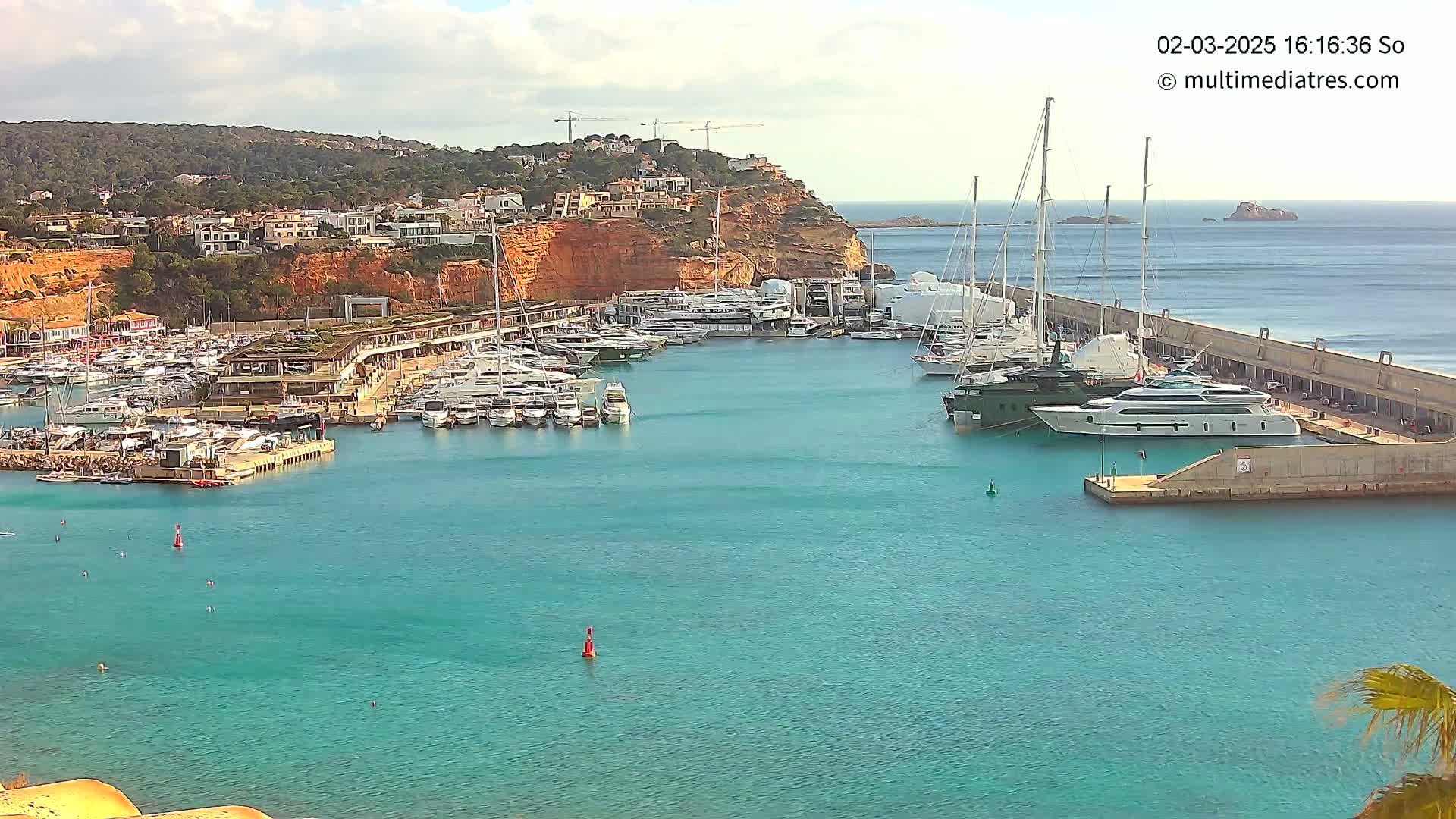 A marina filled with numerous yachts and sailboats is situated along a rocky coast under a partly cloudy sky.