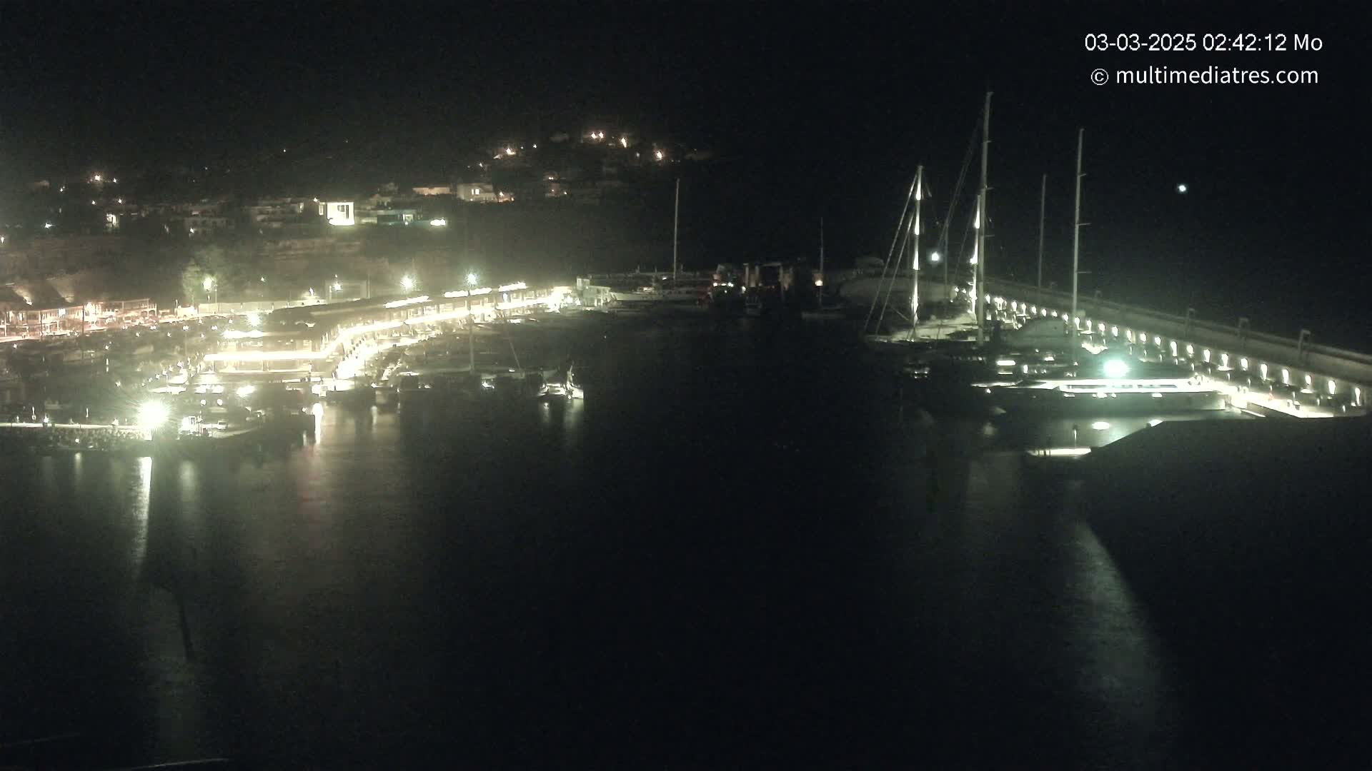 A nighttime view of a marina with numerous boats docked, illuminated buildings along the waterfront, and a dark, calm body of water.