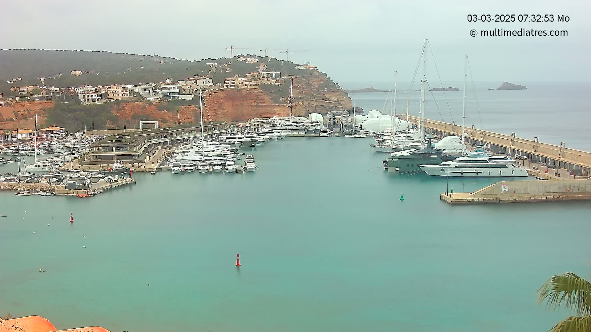 A marina filled with numerous yachts and boats is nestled beside a hillside of buildings under an overcast sky.