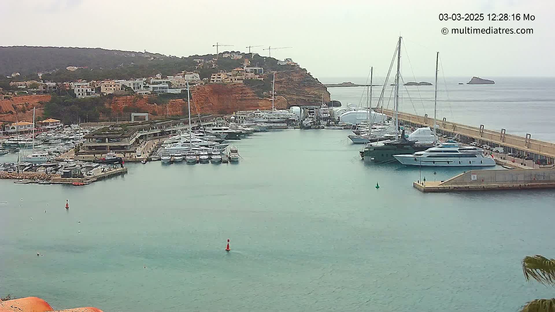 A marina filled with numerous yachts and boats is nestled along a red-cliffed coast under an overcast sky.