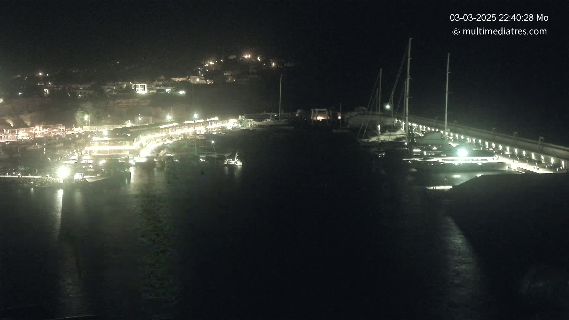 A nighttime, aerial view of a marina filled with boats, illuminated by lights, with a town visible in the background on a hill.