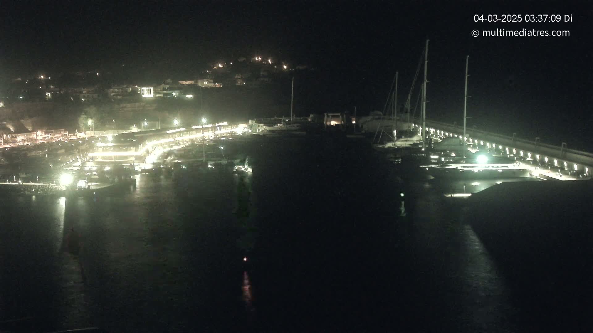 A nighttime aerial view shows a marina filled with boats, docked along a lit seawall, with a town illuminated on a hillside in the background.