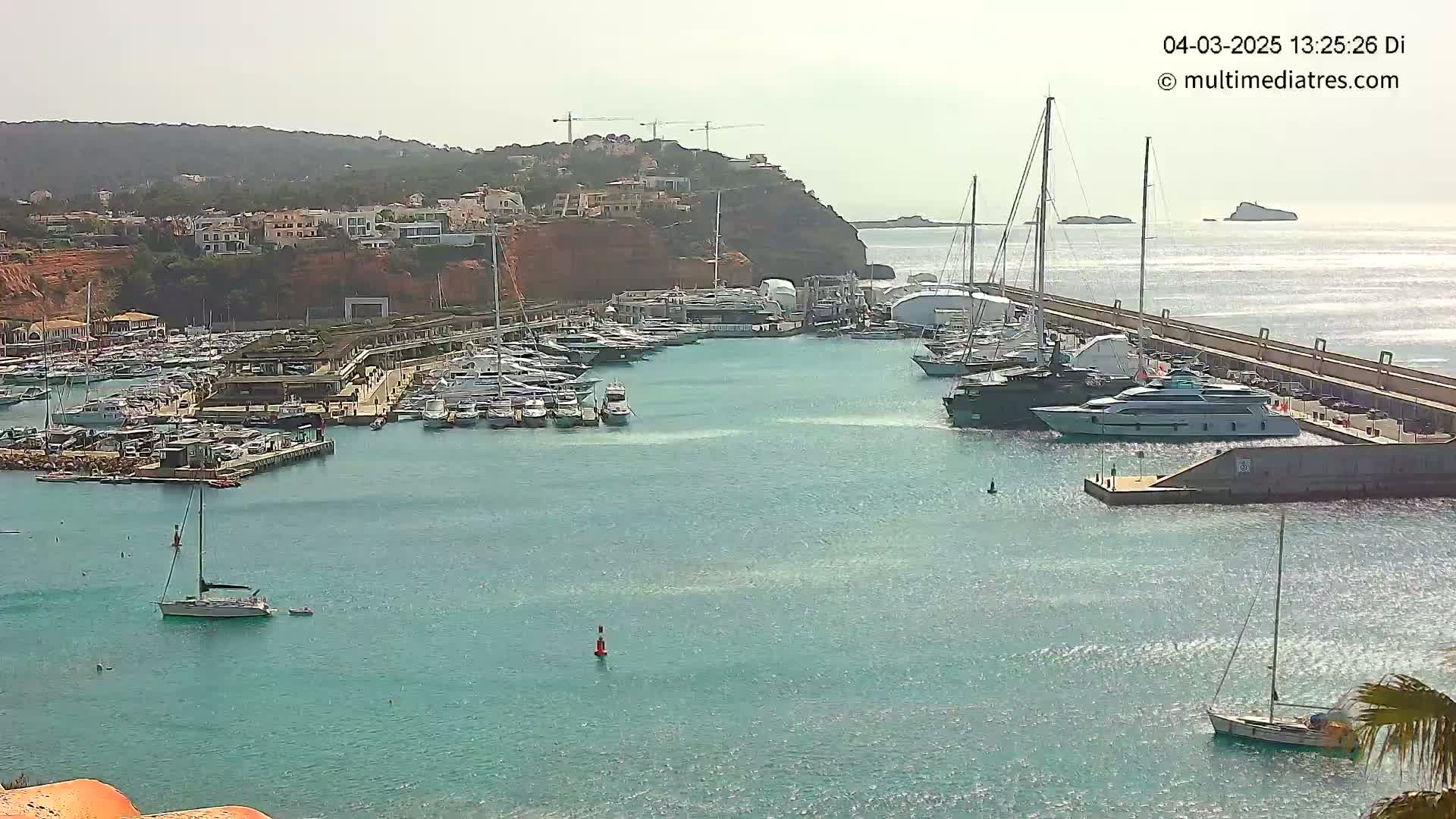 A marina filled with numerous yachts and sailboats is situated near a hillside of buildings under a mostly clear sky.