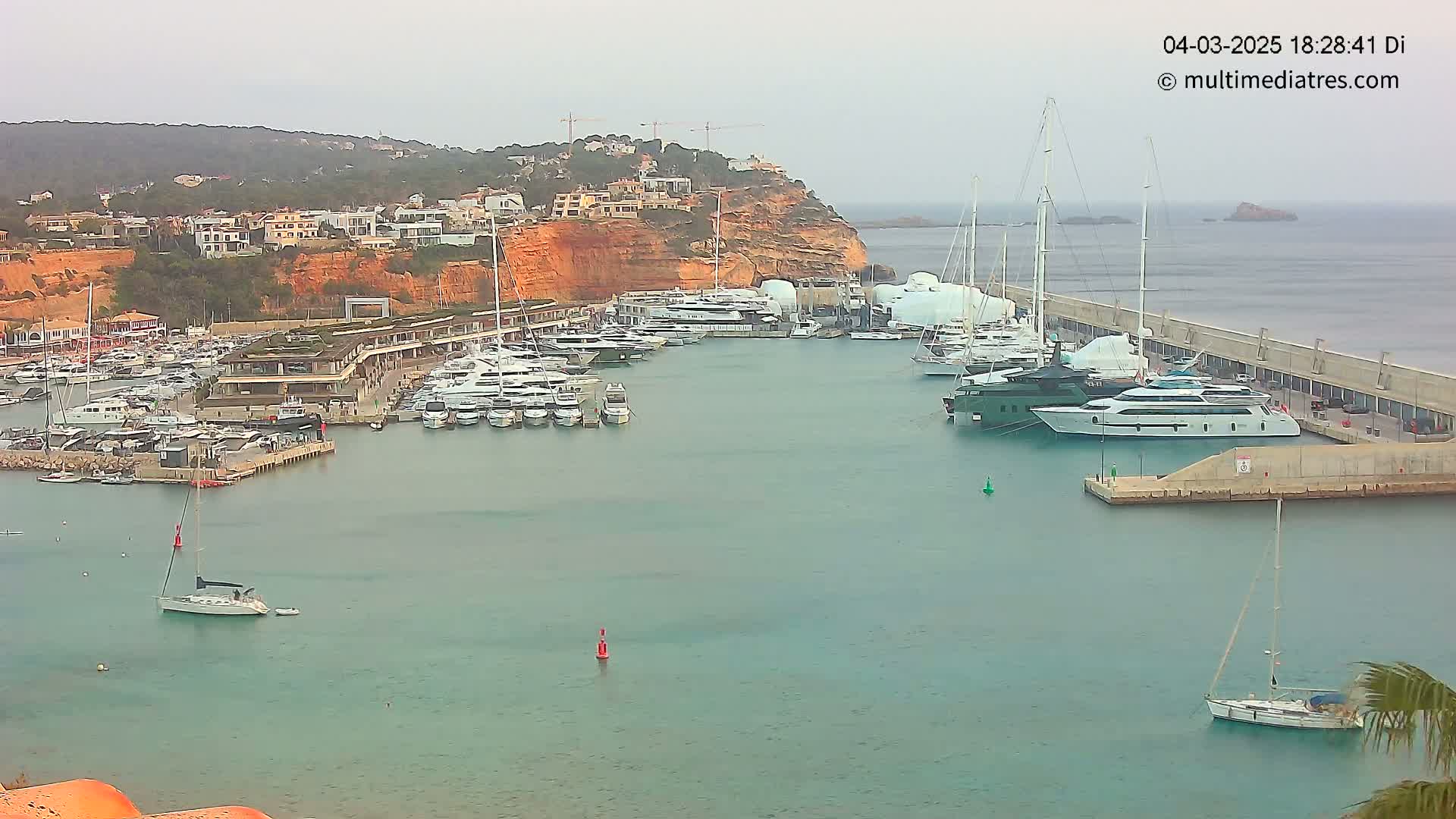 A marina filled with numerous yachts and sailboats is nestled against a hillside of buildings under an overcast sky.