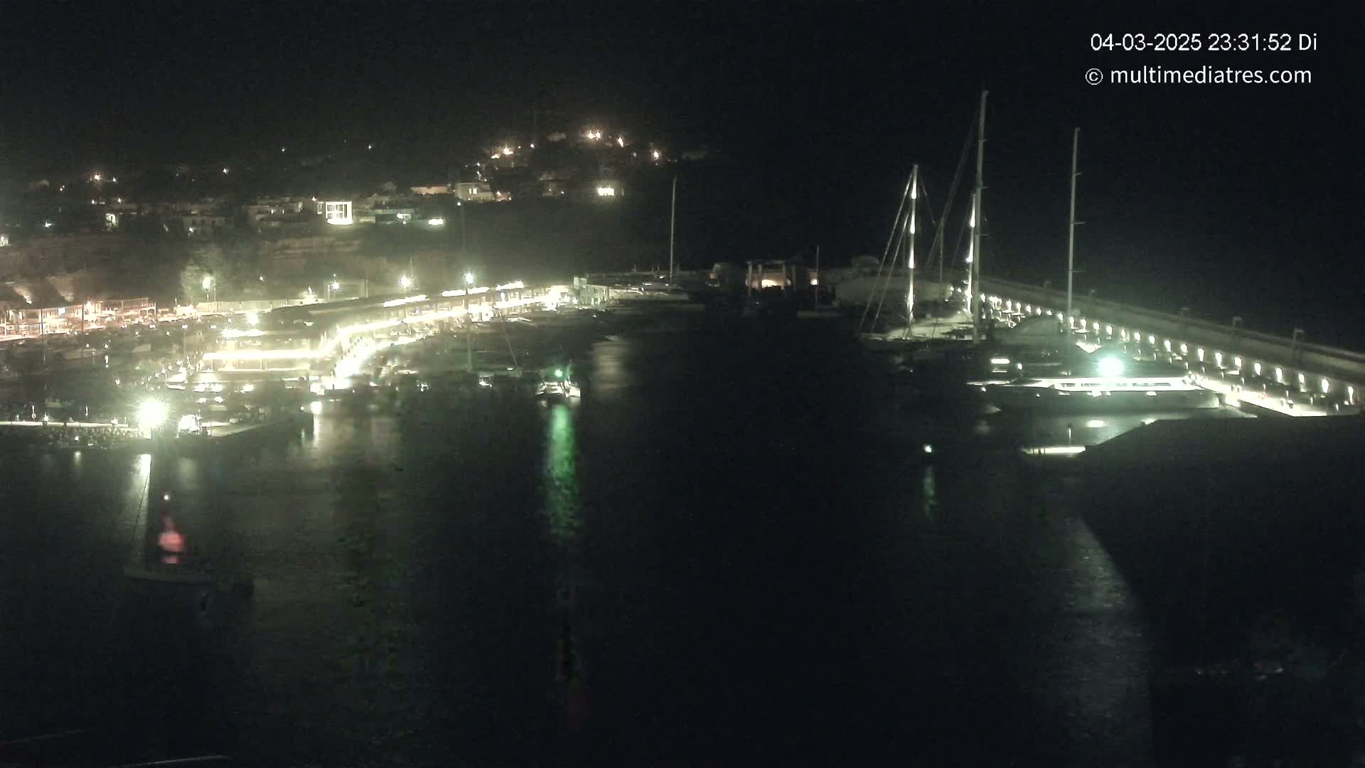 A nighttime, aerial view of a harbor filled with boats, docked along a lit pier, with a hilltop town visible in the background.