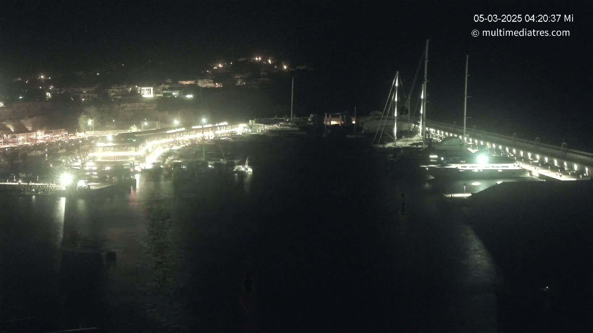 A nighttime view of a harbor filled with boats, illuminated by lights on the docks and buildings along the shoreline.