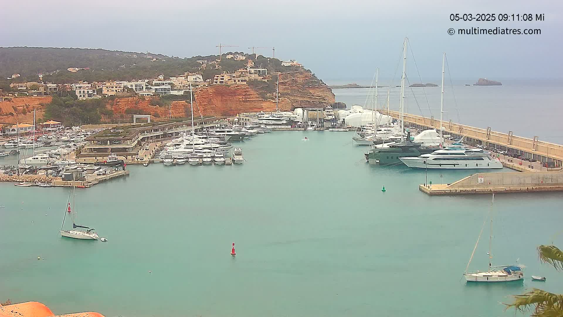 A marina filled with numerous yachts and sailboats is nestled beside a hillside of buildings under an overcast sky.