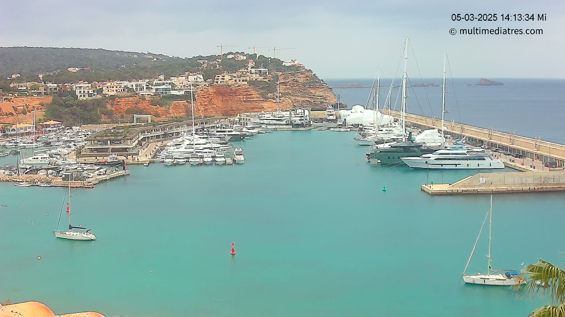 A marina filled with yachts and sailboats is nestled against a rocky cliffside under an overcast sky.