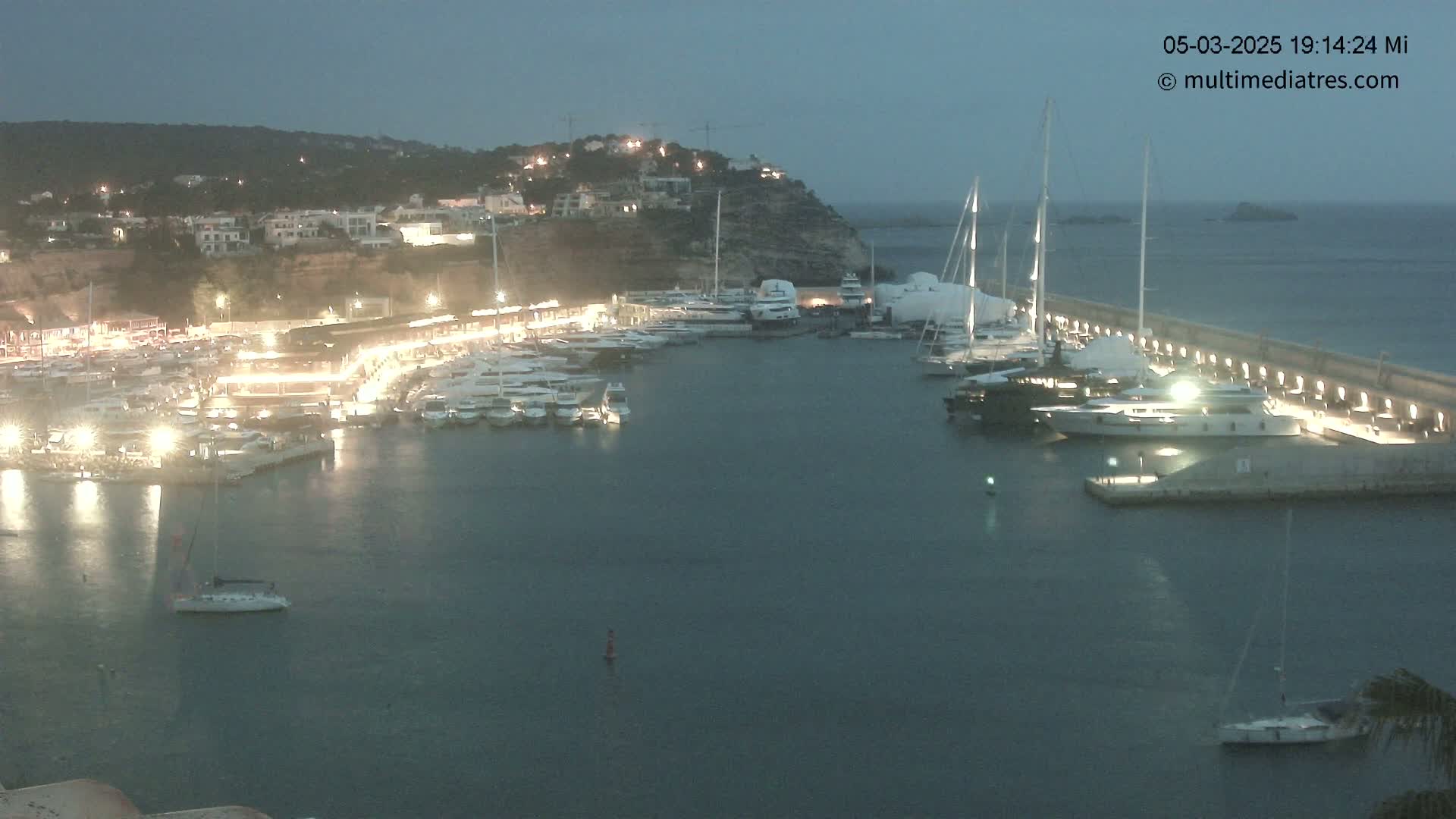 A harbor at night, filled with many yachts and boats docked along a lit seawall under a dark, twilight sky.