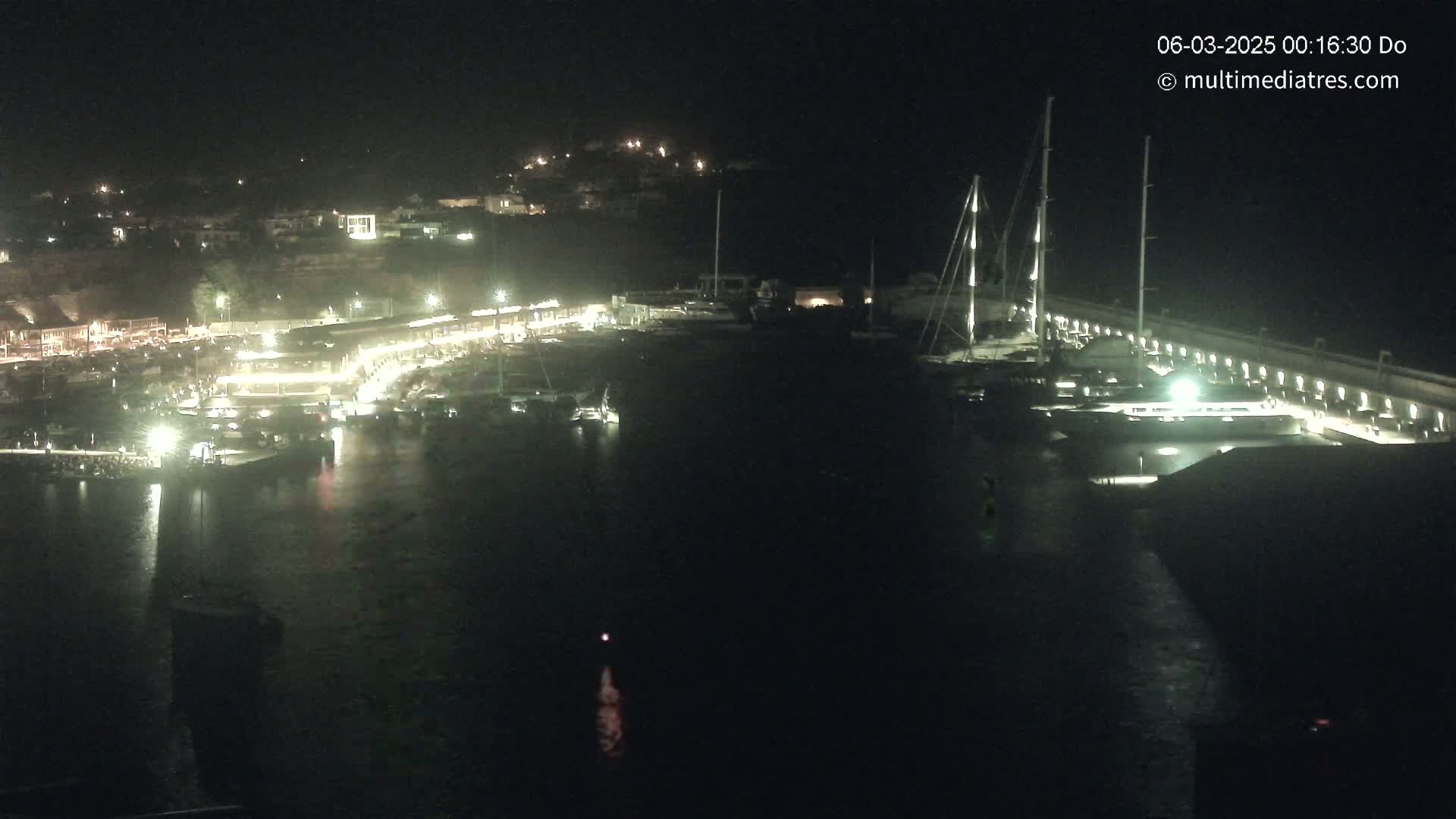 A nighttime aerial view of a harbor with boats docked, illuminated buildings along the shoreline, and hills in the background.