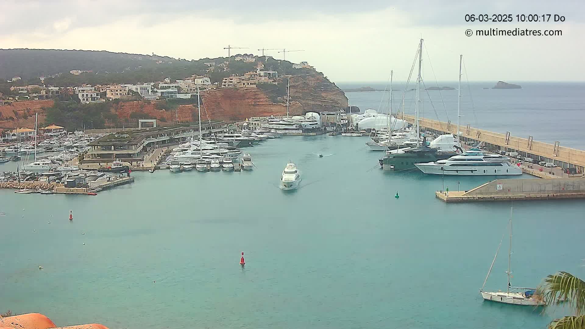 A marina filled with numerous yachts and sailboats is situated on calm, teal water, nestled against a hillside of buildings under an overcast sky.