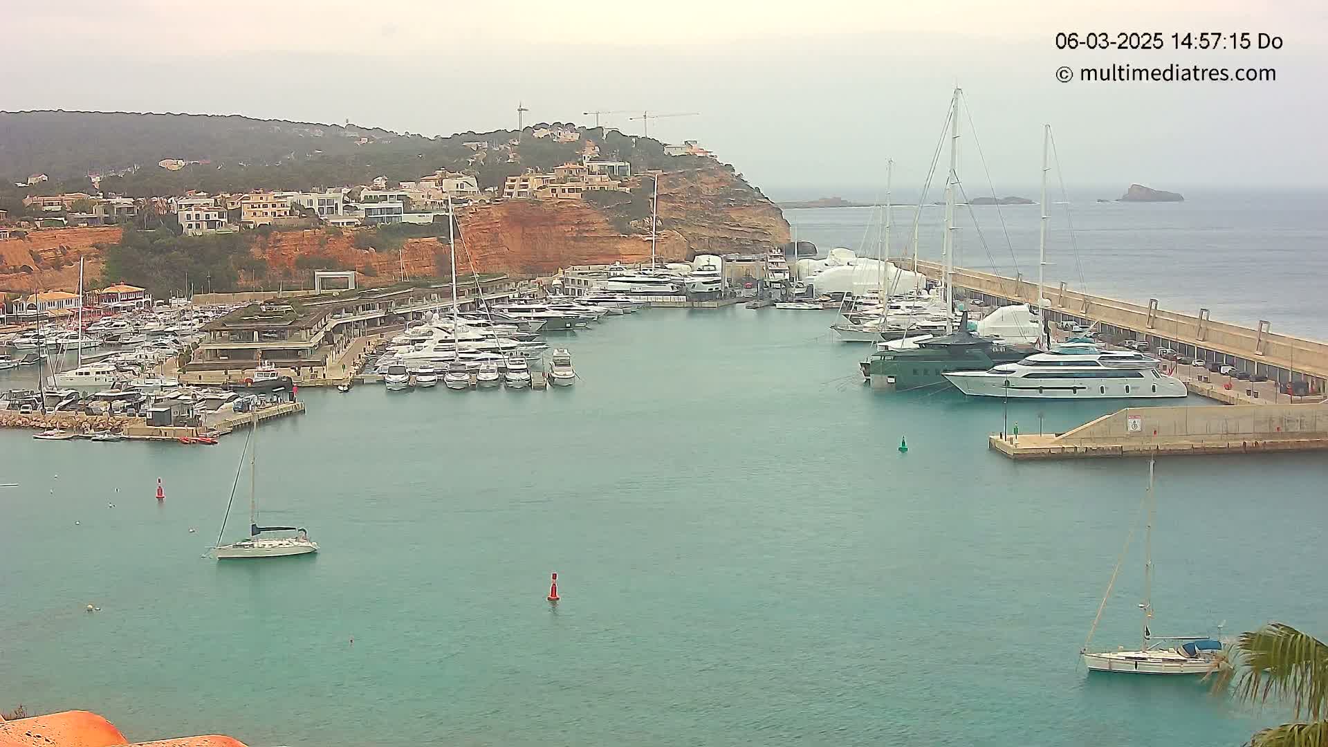 A marina filled with numerous yachts and sailboats is situated next to a hillside of buildings under an overcast sky.