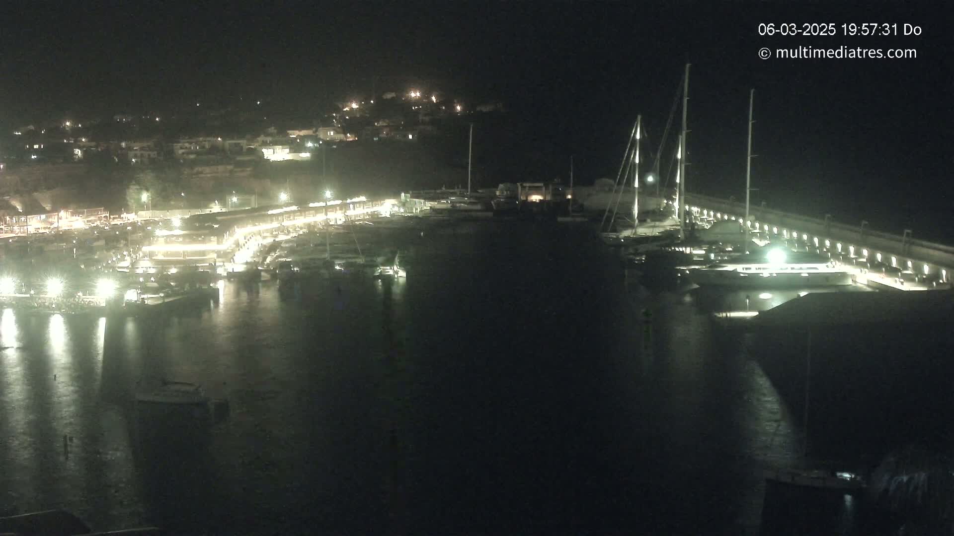 A nighttime view of a marina filled with boats, illuminated by lights against a dark, calm sea.