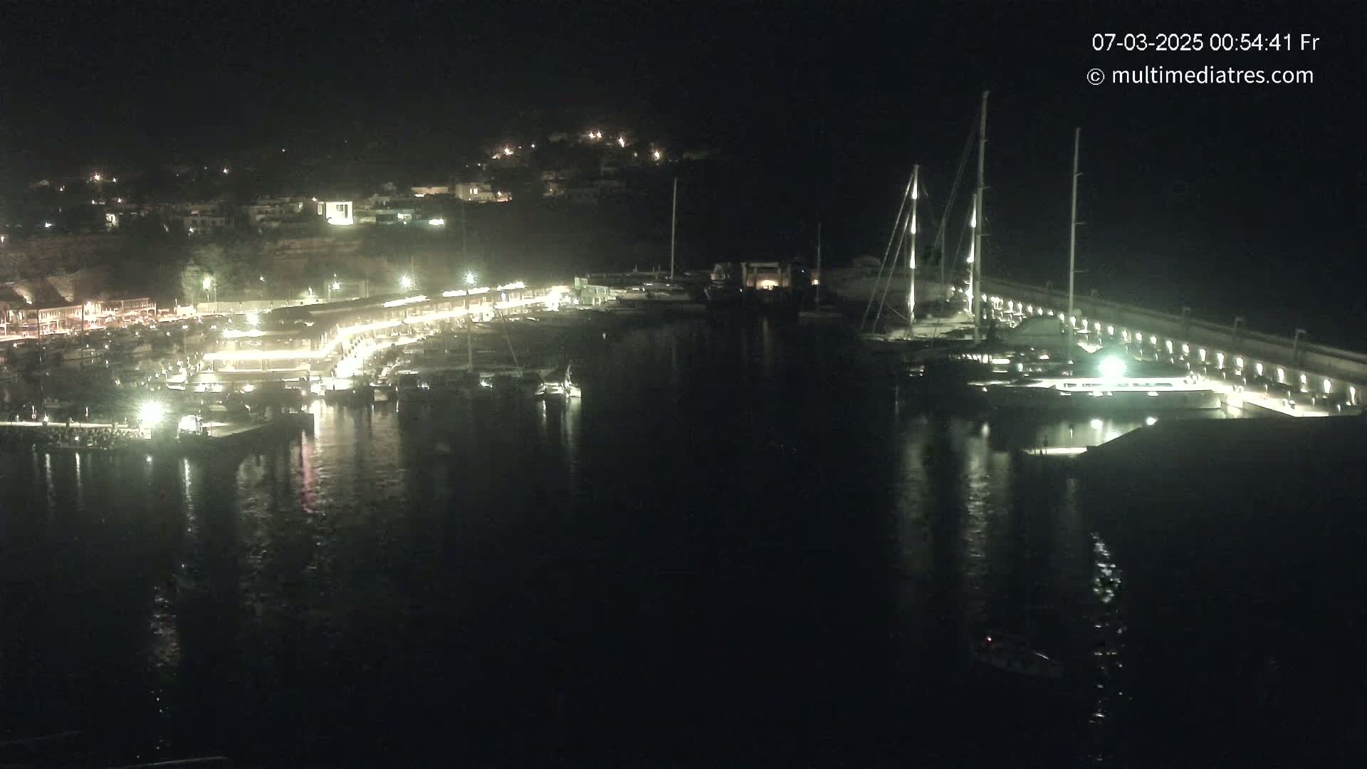 A nighttime view of a marina with boats docked, buildings lit up along the shore, and hills in the background under a clear, dark sky.