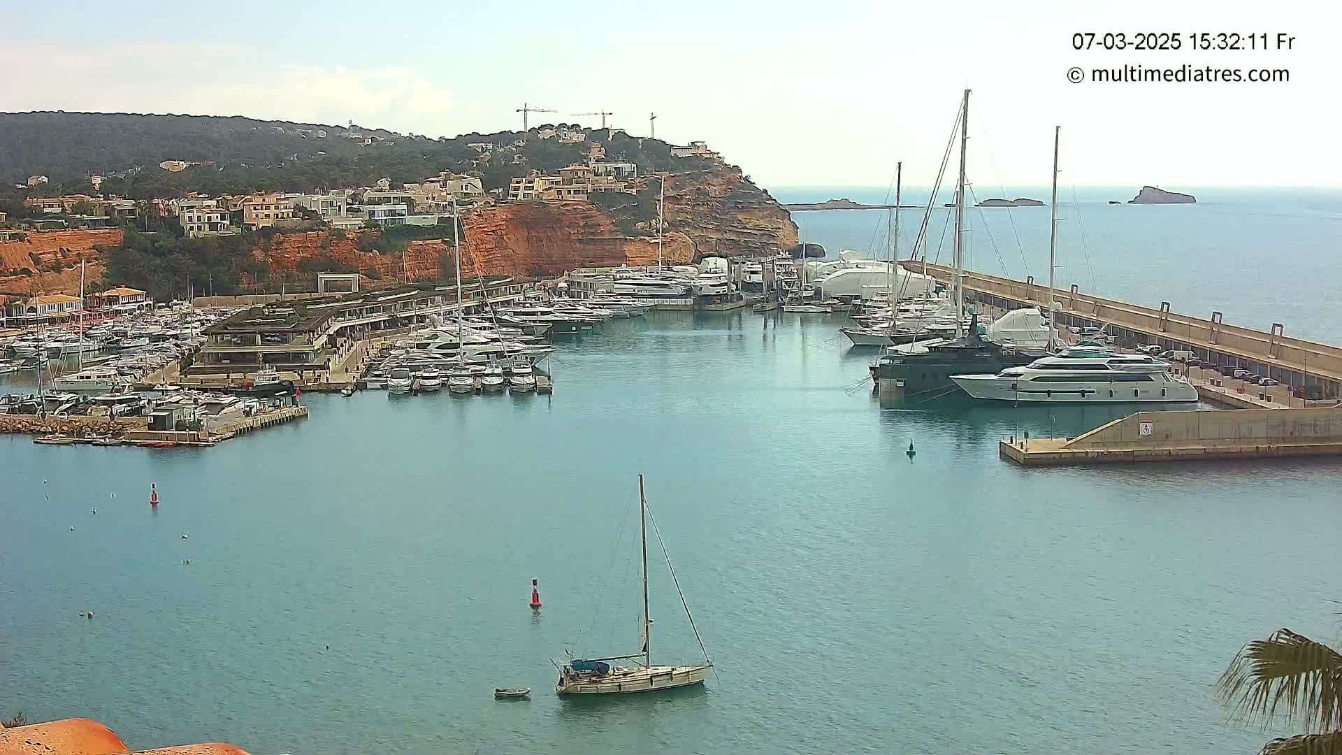 A marina filled with yachts and sailboats is nestled beside a red-cliff hillside under a partly cloudy sky.