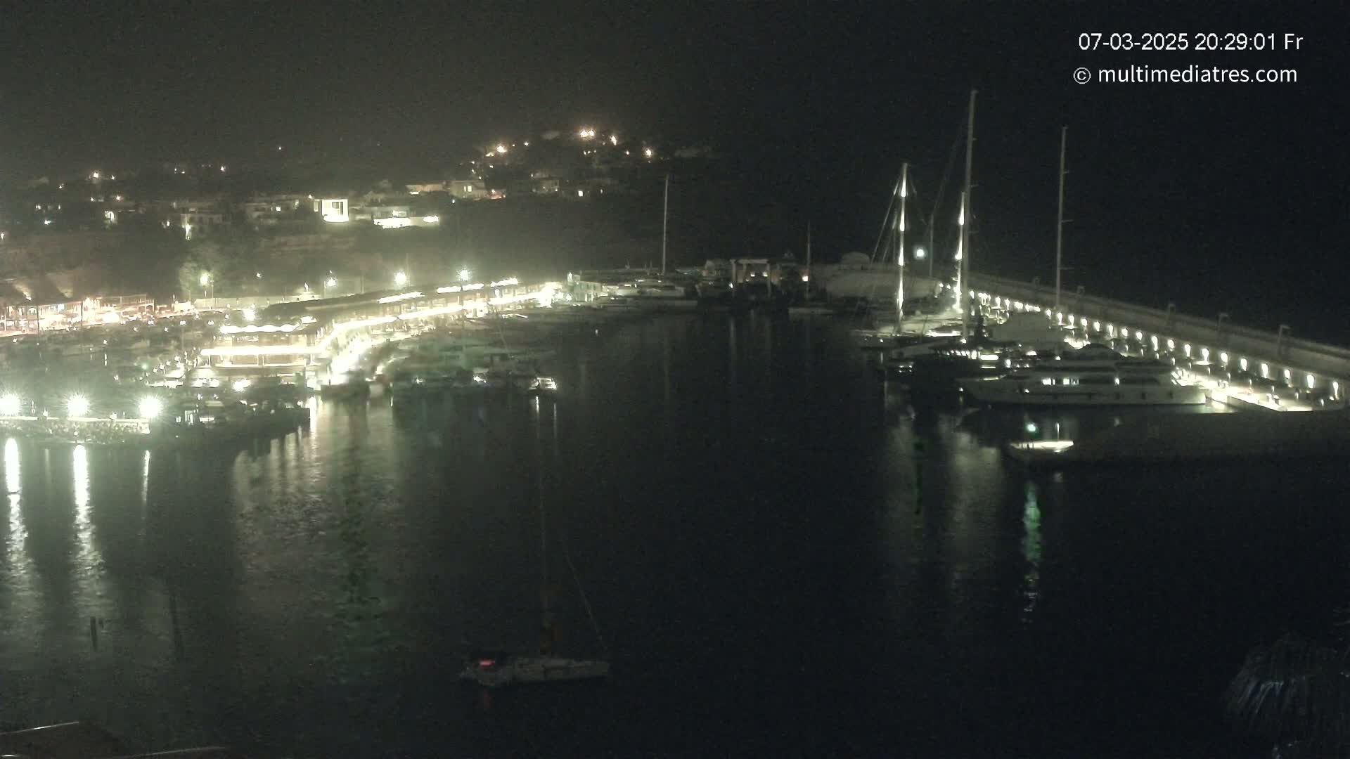 A nighttime view of a marina filled with boats, illuminated by lights along a waterfront, under a dark sky.