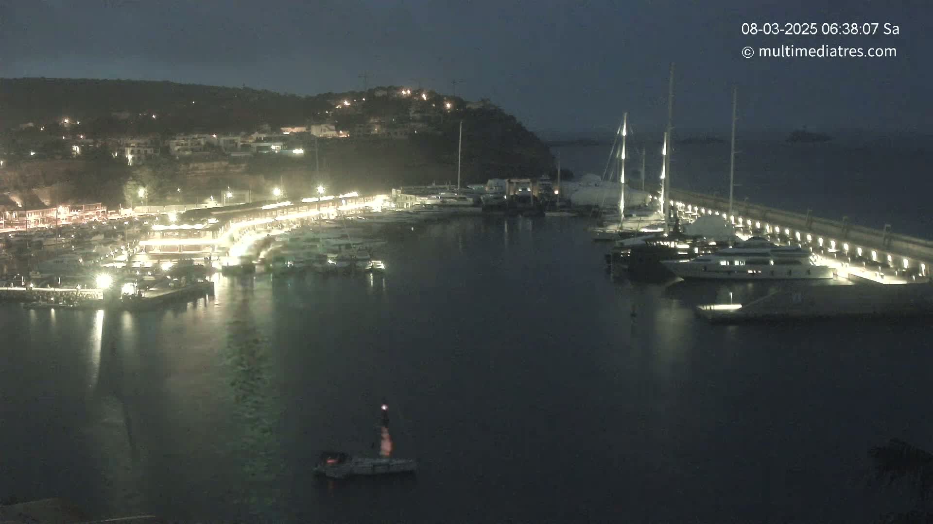 A nighttime view of a marina filled with boats, docked alongside a lit waterfront against a dark hillside.