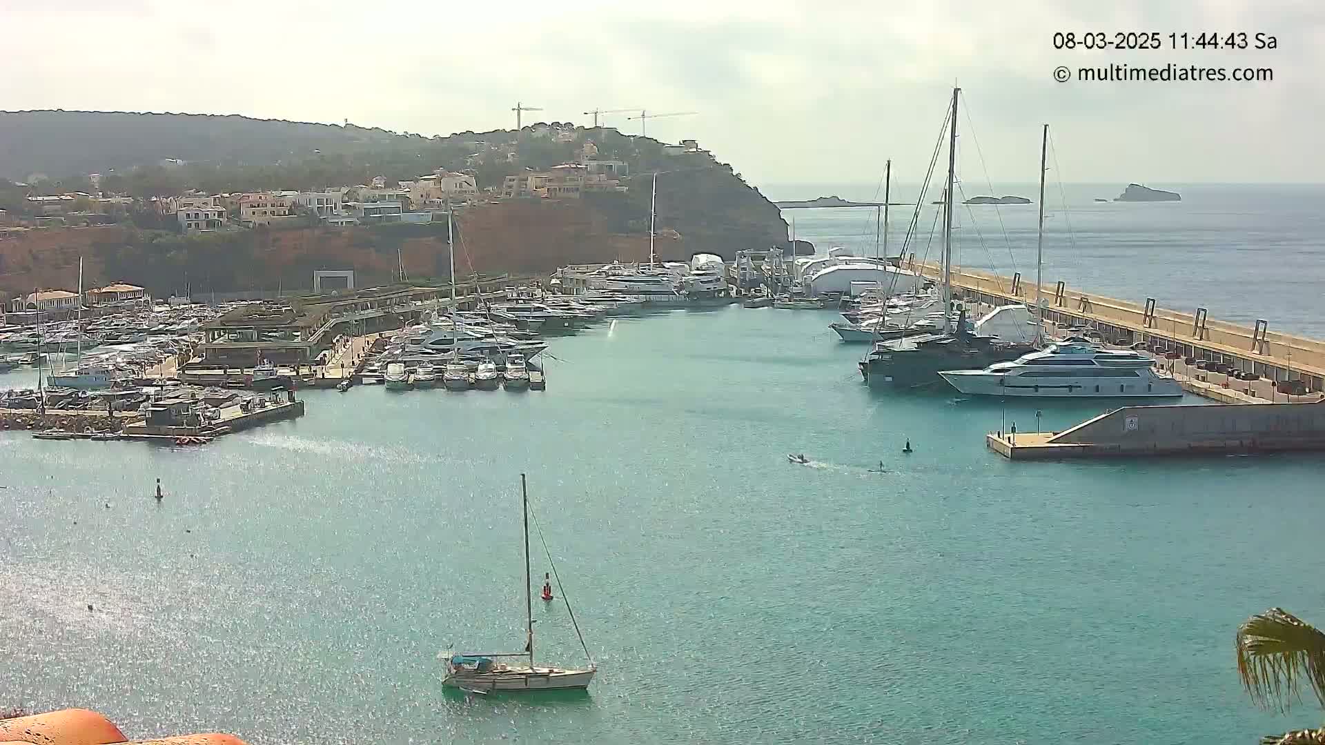 A calm, sunny day shows a marina filled with yachts and sailboats, nestled next to a hillside of buildings, with small islands visible in the distance.