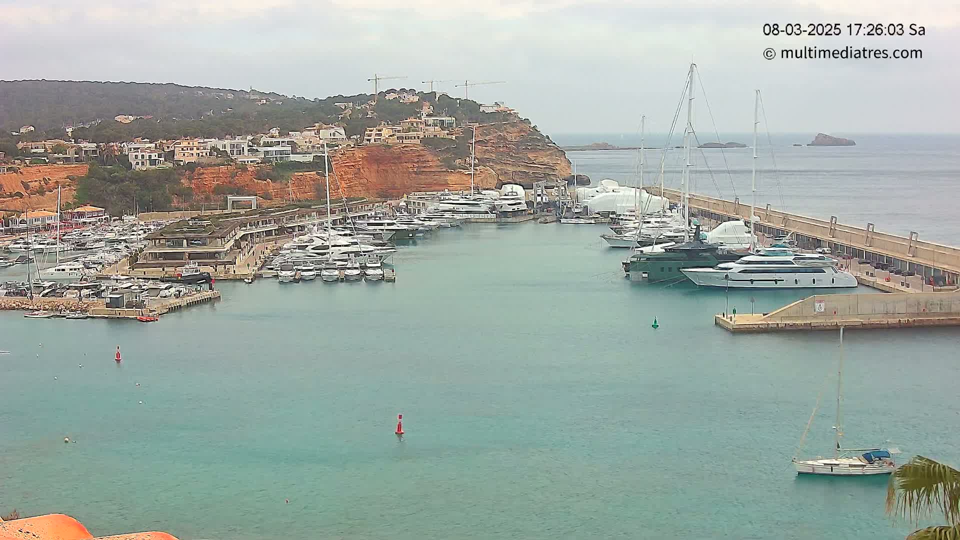 A marina filled with numerous yachts and sailboats is situated next to a hillside town under an overcast sky.